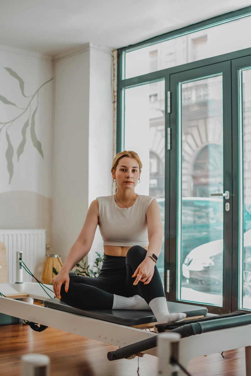 Nő felrakott lábakkal egy pilates gépen pihen és a kamerába néz belvárosi stúdióban