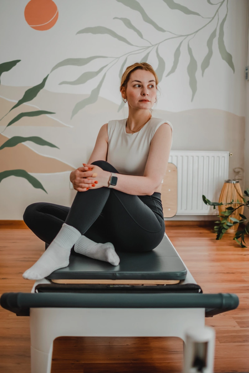 Nő egy pilates gépen felhúzott lábakkal ülve pihen, és az utcára néző ablakon néz ki