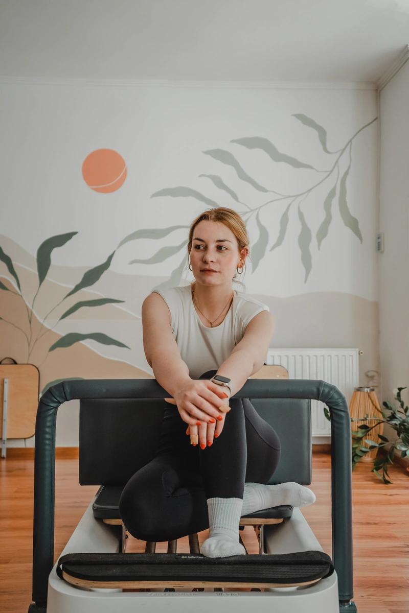 Nő a pilates gépen felhúzott lábakkal ül, kezeit a korlátra rakva oldalra néz