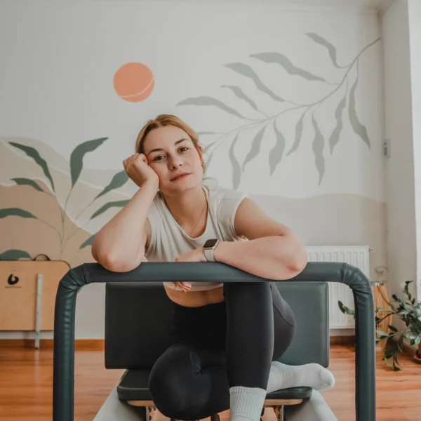 Nő a pilates gépen felhúzott lábakkal ül, arcát egy kezével, oldalt támasztja meg a gép korlátján és a kamerába néz