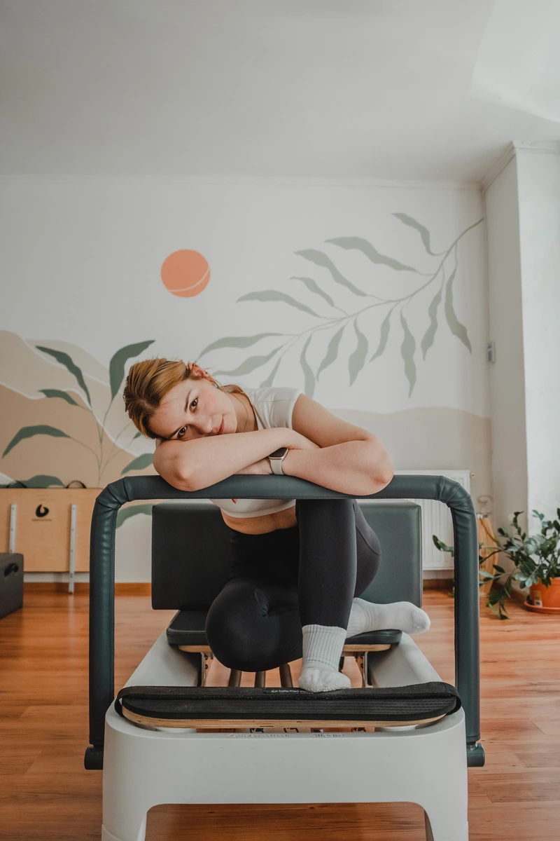 Nő a pilates gépen felhúzott lábakkal ül, karjait a pilates gép korlátján összefonja, és oldalra dönti a fejét