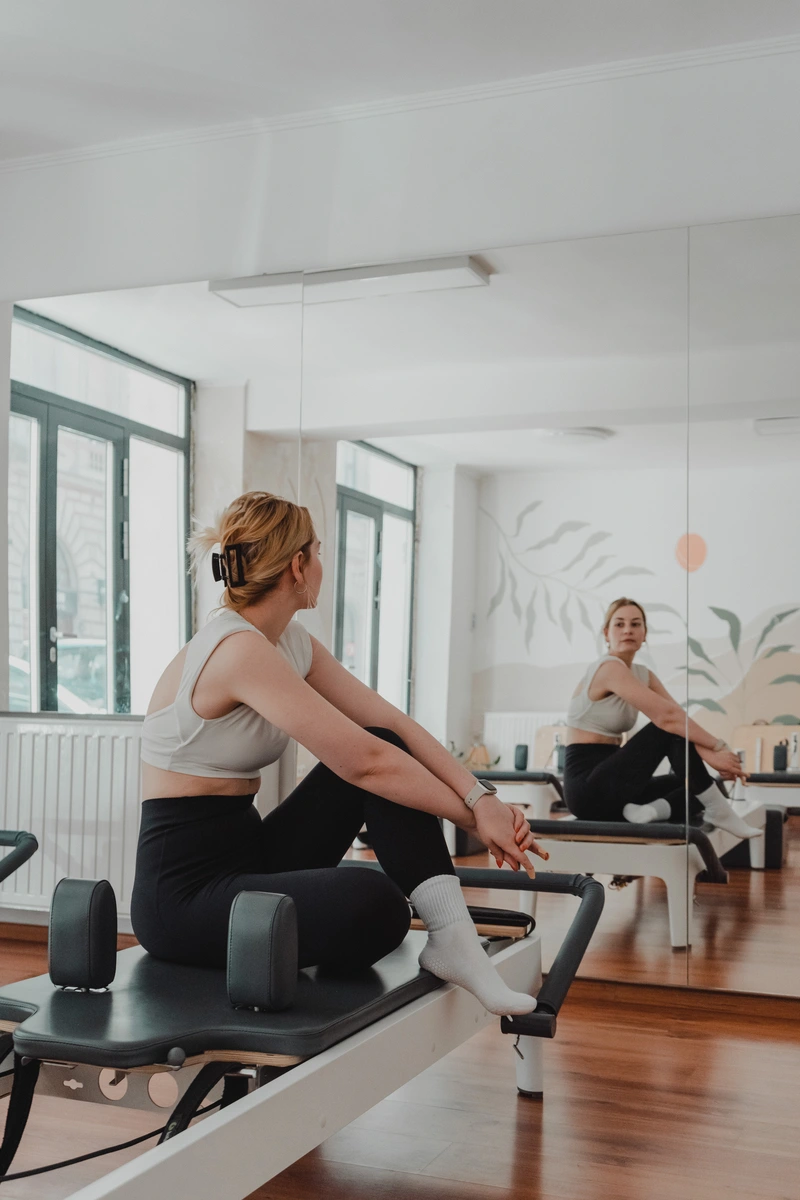 Nő oldalra fordított fejjel nézi magát a tükörben egy pilates gépen ülve
