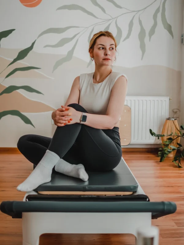 Nő egy pilates gépen felhúzott lábakkal ülve pihen, és az utcára néző ablakon néz ki
